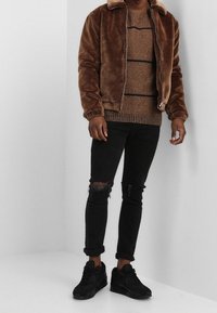 Brun fuskpälsjacka med krage, stickad brun tröja med mörka ränder, svarta slitna jeans och svarta sneakers.