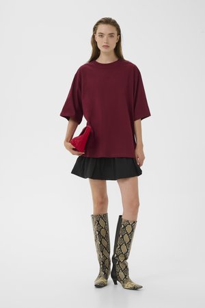 Žena u širokoj bordo košulji, crnoj mini suknji i čizmama do koljena od zmijske kože, drži crvenu torbicu na bijeloj pozadini.