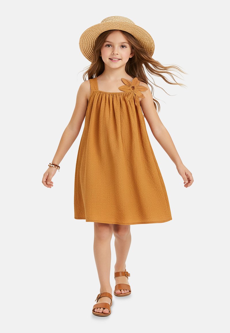 Jeune fille marchant dans une robe moutarde texturée avec un détail de fleur, portant un chapeau de paille, un bracelet et des sandales marron sur fond blanc.