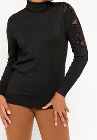 Svart polotröja med glittrig textur, med utskärningar på ärmarna. Kombinerad med bruna byxor.