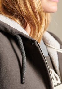 Sudadera con capucha gris oscuro, con un suave forro de felpa, presenta una cremallera, cordones texturizados y un logo bordado en la parte delantera.