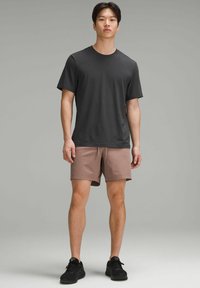 Schwarzes Kurzarm-T-Shirt, lockere Passform, kombiniert mit hellbraunen Shorts mit Kordelzugtalie. Schwarze Sport-Schuhe runden das Outfit ab.