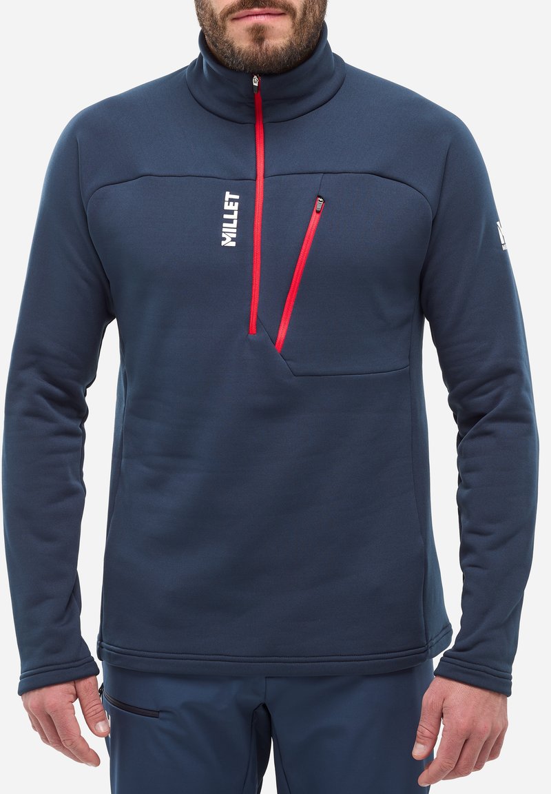 Pull-over en polaire marine avec un col haut, possède une petite poche poitrine zippée et des accents rouges. Texture douce, design ajusté et branding minimal.
