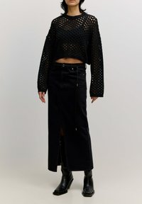 Zwarte gebreide cropped sweater met open geweven patroon, gecombineerd met een zwarte denim maxi rok met een zijsplit en trekkoorddetail. Zwarte enkellaarsjes.