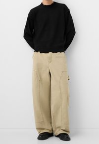 Svart tröja med ribbat halsringning och fransigt släpp, parat med beige vida byxor med tryckknappsfickor. Svarta skor fulländar outfiten.