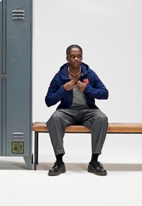 Marineblå hettegenser med glidelås foran og logo, grå bukser og svarte sko. Benk å sitte på ved siden av et metallskap med logodesign.