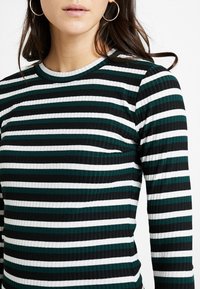 Gestreiftes Langarmshirt mit horizontalen Streifen in Dunkelgrün, Schwarz und Weiß. Rippstruktur mit Rundhalsausschnitt.