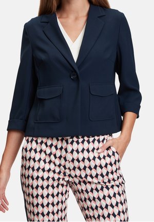 Donna che indossa un blazer blu navy con tasche a pattina, una maglietta bianca con scollo a V e pantaloni a fantasia con forme geometriche blu navy e rosa.