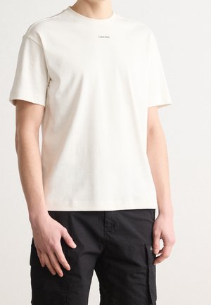 Man draagt een eenvoudige witte Calvin Klein T-shirt en zwarte cargobroek, staand tegen een effen witte achtergrond.