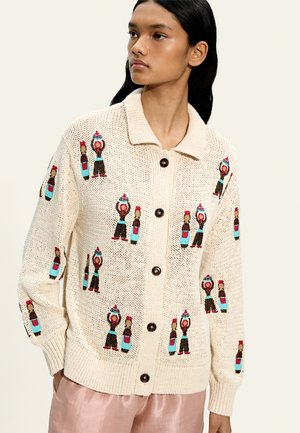 Młoda kobieta ubrana w kremowy sweter na guziki z kolorowymi haftowanymi postaciami i butelkami, w połączeniu z jasnoróżowymi spodniami.