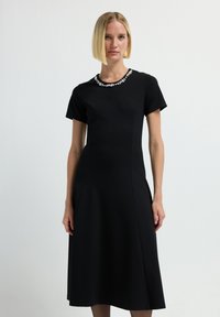 Vestido negro con mangas cortas y falda acampanada, con un detalle texturizado en el cuello. Tejido liso y corpiño ajustado.