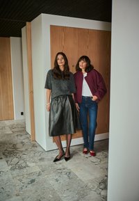 Twee vrouwen staan naast elkaar. De een draagt een grijze gebreide top en een zwarte leren rok; de ander draagt een bordeauxrode bomberjack en blauwe jeans.