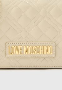 Μπεζ δερμάτινη θήκη με καπιτονέ σχέδιο και χρυσό λογότυπο "LOVE MOSCHINO". Διαθέτει διακοσμητικές ραφές σταυρωτού τύπου και απαλή υφή. Συμπαγής σχεδιασμός.
