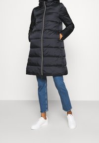 Schwarzer Steppmantel mit Kapuze, Reißverschluss und Seitentaschen. Verfügt über ein gestepptes Design und kontrastierende Stoffärmel. Getragen über Jeans und Sneakers.