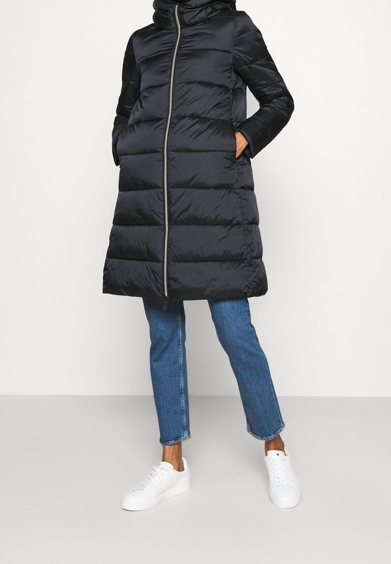 Schwarzer Steppmantel mit Kapuze, Reißverschluss und Seitentaschen. Verfügt über ein gestepptes Design und kontrastierende Stoffärmel. Getragen über Jeans und Sneakers.