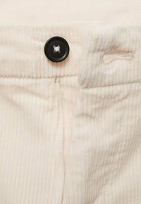 Bouton noir fixé sur un tissu en velours côtelé crème avec des détails visibles de boutonnière et de couture.