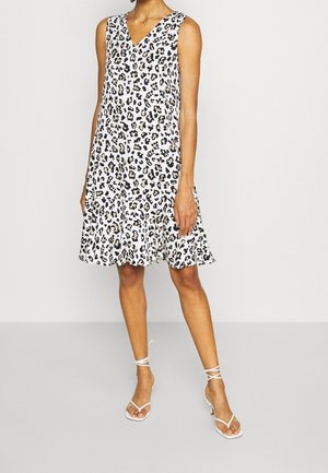 Femme portant une robe blanche sans manches avec un imprimé léopard noir et beige et des sandales blanches à talons avec brides, debout devant un fond uni.