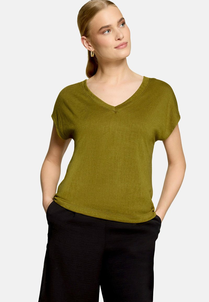 Femme aux cheveux blonds attachés en queue de cheval, portant des boucles d'oreilles anneaux dorés, un haut vert olive à col en V et manches courtes, et un pantalon noir, les mains dans les poches.