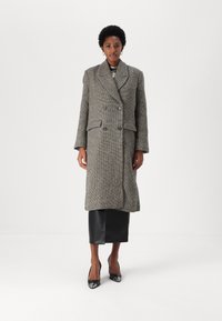 Abrigo de espiga en gris y negro, de doble botonadura, con grandes solapas, bolsillos laterales y un diseño de largo medio, llevado sobre una falda de cuero negra.