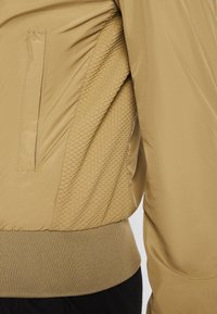 Khaki Bomberjacke mit glatter Stoffoberfläche, strukturiertem Seitenpanel, geripptem Saum und diskretem Seitentasche.