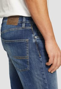 Blauwe denim jeans met een gestructureerde oppervlakte, voorzien van een bruine leren label op de tailleband en gele stiksels langs de zakken.