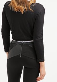 Donna che indossa un top nero a maniche lunghe e pantaloni neri con una cerniera centrale sulla schiena e pannelli in pelle sui fianchi, in piedi davanti a uno sfondo bianco.