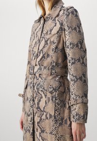 Manteau à motif peau de serpent en beige et noir, avec une taille ceinturée, une fermeture à boutons et deux poches avant avec rabats. Tissu texturé.