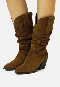 Botas de tobillo de ante marrón con un diseño arrugado, punta afilada y tacón apilado. Presenta detalles de costura a lo largo de los lados.