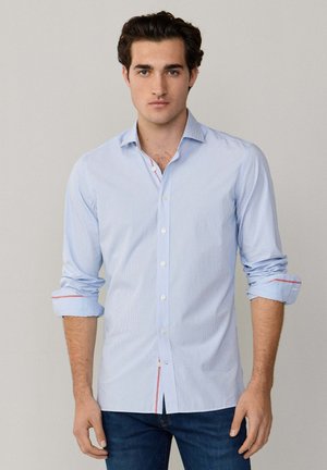 Junger Mann trägt ein hellblaues Hemd mit hochgekrempelten Ärmeln und dunkelblaue Jeans und steht vor einem einfarbigen hellen Hintergrund.