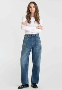 Jeans en denim bleu taille haute avec une coupe décontractée, dotés de grandes poches avant et d'une texture légèrement décolorée. Associés à un haut blanc à manches longues.