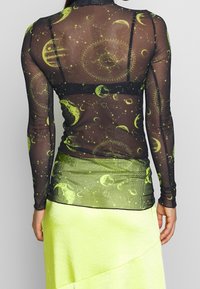Mustat pitkät hihat -paita, jossa on läpinäkyvää materiaalia ja vihreitä kosmisia kuvioita. Yhdistetty neonkeltaisen hameen kanssa, joka korostaa kerrostettua muotoilua.