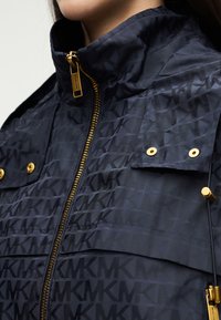 Γυναίκα που φοράει ναυτικό μπουφάν με επαναλαμβανόμενο μοτίβο "MK", χρυστό φερμουάρ και κουμπιά στα τσέπες στο στήθος, εμφανιζόμενη από το λαιμό μέχρι το μέσο του κορμού.