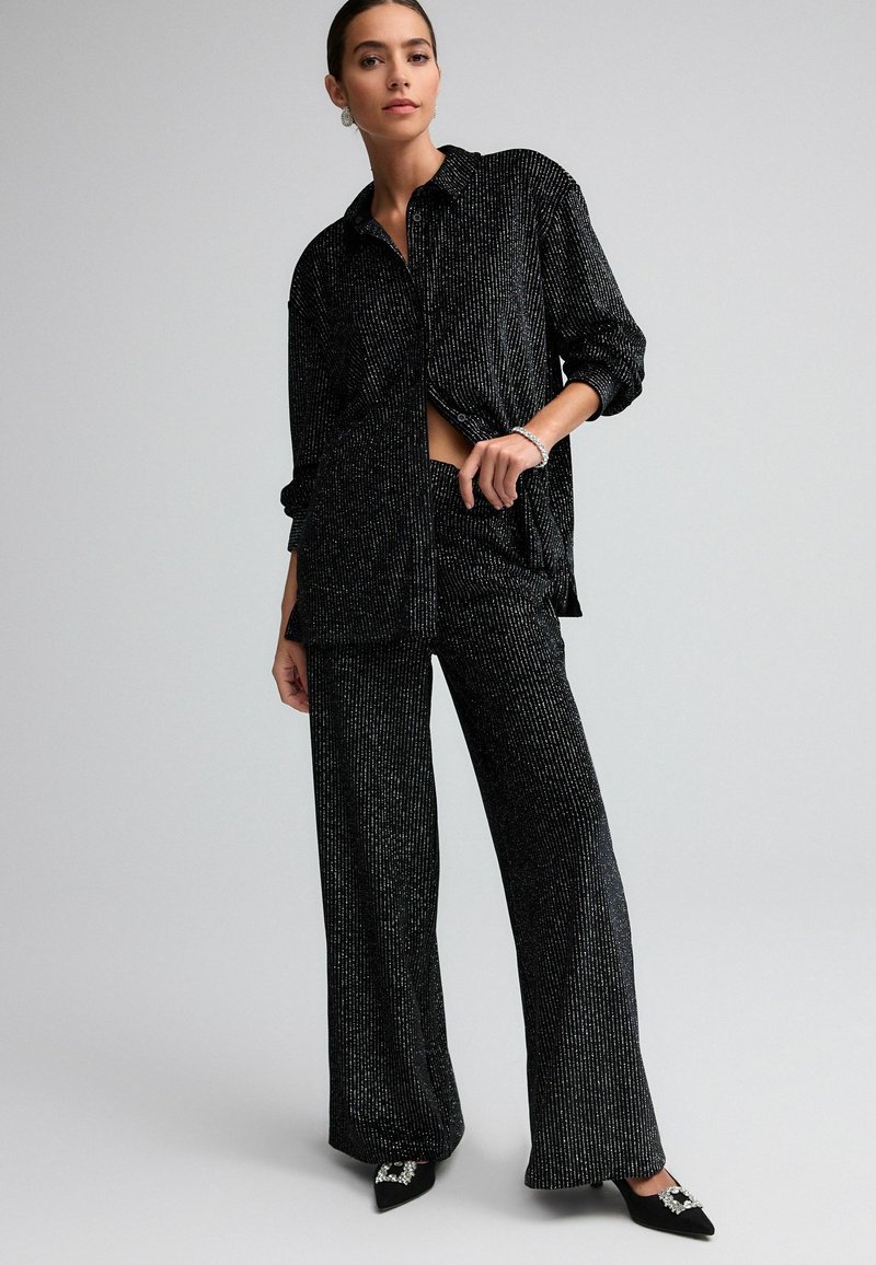 Black, shiny two-piece outfit with a loose collared shirt and wide-leg pants, featuring vertical metallic stripes and pointed-toe black shoes.