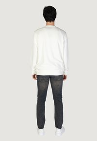 Camicia bianca a maniche lunghe con polsini a coste, abbinata a jeans scuri e scarpe da ginnastica bianche, vista da dietro. Design semplice, senza loghi visibili.