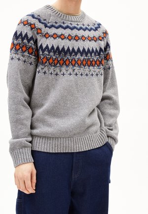 Person iført en grå striktrøje med orange, navy og sorte geometriske mønstre over brystet og skuldrene, kombineret med mørkeblå bukser.