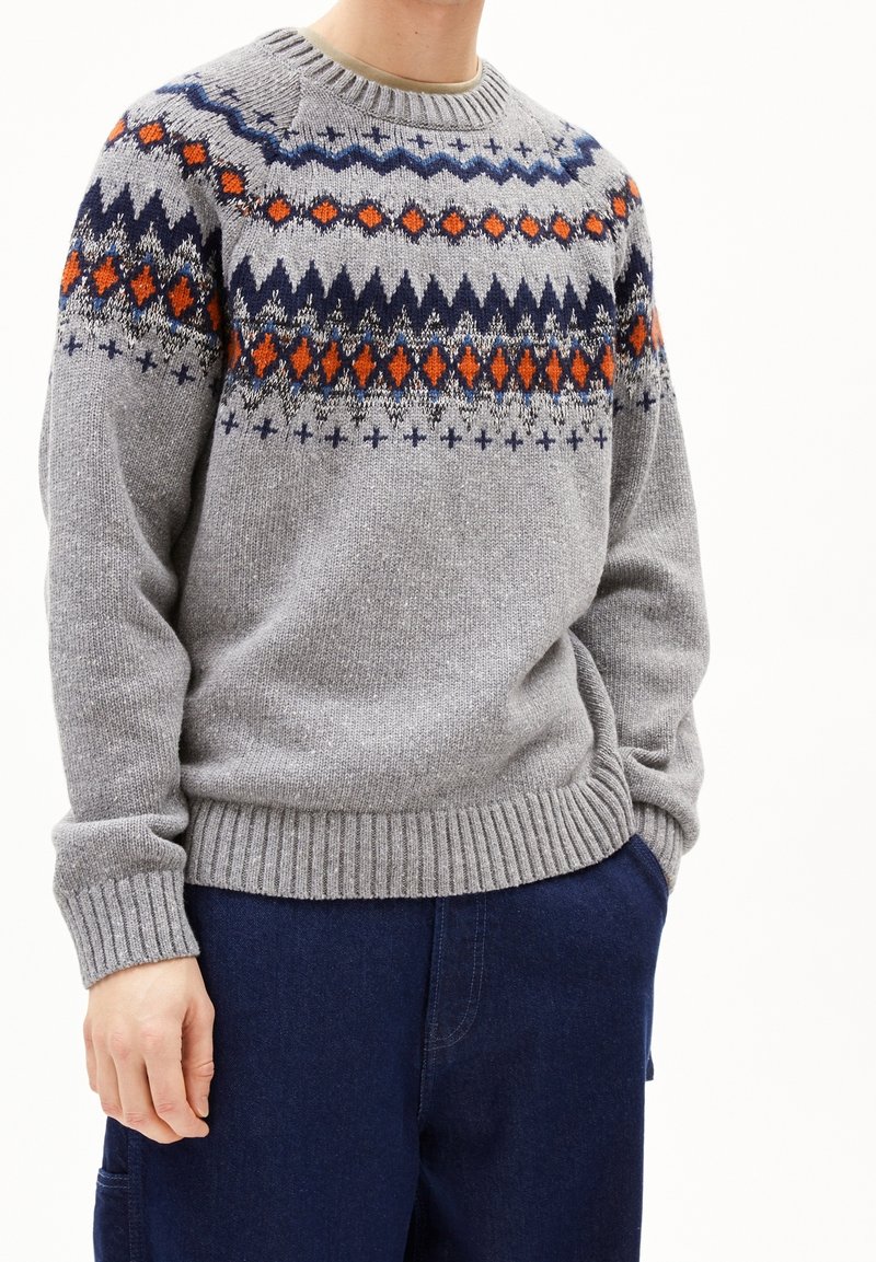 Personne portant un pull en maille gris avec des motifs géométriques orange, marine et noirs sur la poitrine et les épaules, associé à un pantalon bleu foncé.