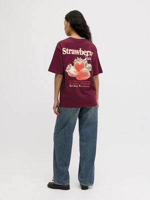 Femme portant un t-shirt bordeaux avec un grand motif fraise et du texte dans le dos, assorti à un jean bleu ample et des chaussures noires.
