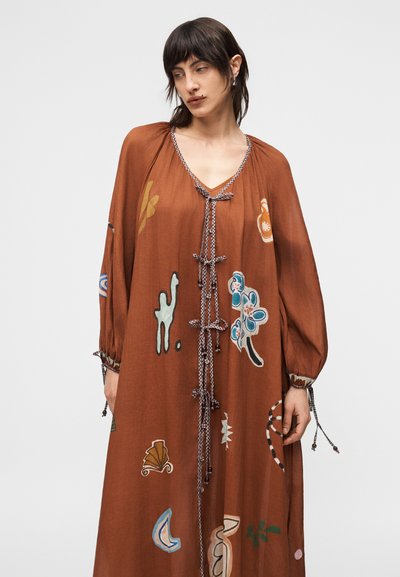 Γυναίκα που φοράει ένα φαρδύ, μακρύ φόρεμα σε σκουρόχρωμο σκουριασμένο χρώμα με πολύχρωμα αφηρημένα λουλουδένια σχέδια και δεμένα κορδόνια στο μπροστινό μέρος και στα μανίκια.