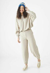Beige sweatsuit met een losse, overgrote sweatshirt met manchetten en bijpassende taps toelopende broek. Draagt een blauwe en crèmekleurige beanie.