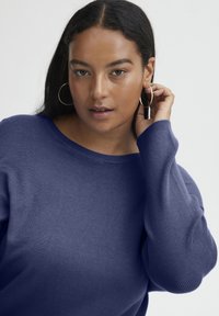 Maglione blu a maglia con scollatura ampia e maniche lunghe. Tessuto liscio con polsini a coste. Modella che indossa orecchini a cerchio e capelli acconciati leggermente.