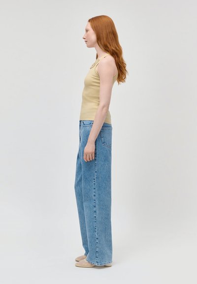 Jeune femme aux longs cheveux roux portant un débardeur beige, un jean bleu à jambes larges et des chaussures beiges, debout de profil sur un fond uni.