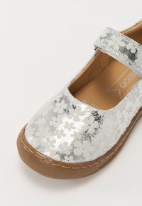 Silberne Mary Jane Schuh mit weißem Blumenmuster, brauner Sohle und Velcro-Riemen auf einem weißen Hintergrund.