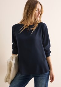 Donkerblauwe top met lange mouwen, een gestructureerd patroon, ronde halslijn en opgerolde mouwen. Het model houdt een crèmekleurige, donzige deken vast.
