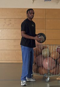 En person i en sort t-shirt og blå jeans holder en basketball nær en trådvogn fyldt med forskellige farvede bolde i et gymnastiklokale.