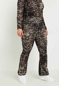 Leopardmönstrad, stretchig velouroutfit med långa ärmar och vida byxor. Färgerna inkluderar nyanser av brunt, svart och beige. Vita sneakers.