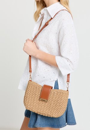 Woman wearing white eyelet blouse and denim skirt holding a woven crossbody bag with brown leather strap and clasp.