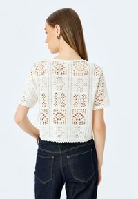 Top de ganchillo blanco con mangas cortas, que presenta un intrincado patrón de calados y un dobladillo ondulado. Combinado con pantalones de mezclilla oscuros.