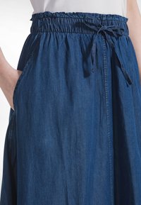 Una falda de mezclilla azul oscuro de longitud media con una pretina elástica con lazada, bolsillos laterales y una tela lisa ligeramente texturizada.