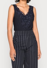 Kvinna klädd i en ärmlös svart topp med blommig spets vid halsen och högmidjade svarta byxor med vita vertikala ränder, med avslappnade händer vid sidorna.