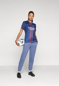 Marinblå fotbollströja med ett rött diagonalt mönster och logotyp, i kombination med ljusblå sweatpants och svarta sneakers. Boll i handen.